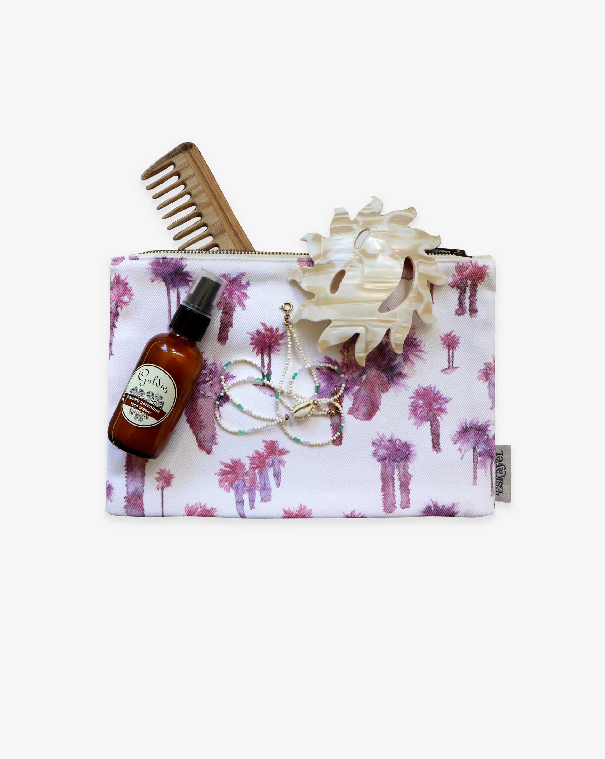 The Perfect Palm Pouch in Persimmon includes a wooden comb, a white hair clip, a bottle of face cream, and a pastel-colored beaded necklace.