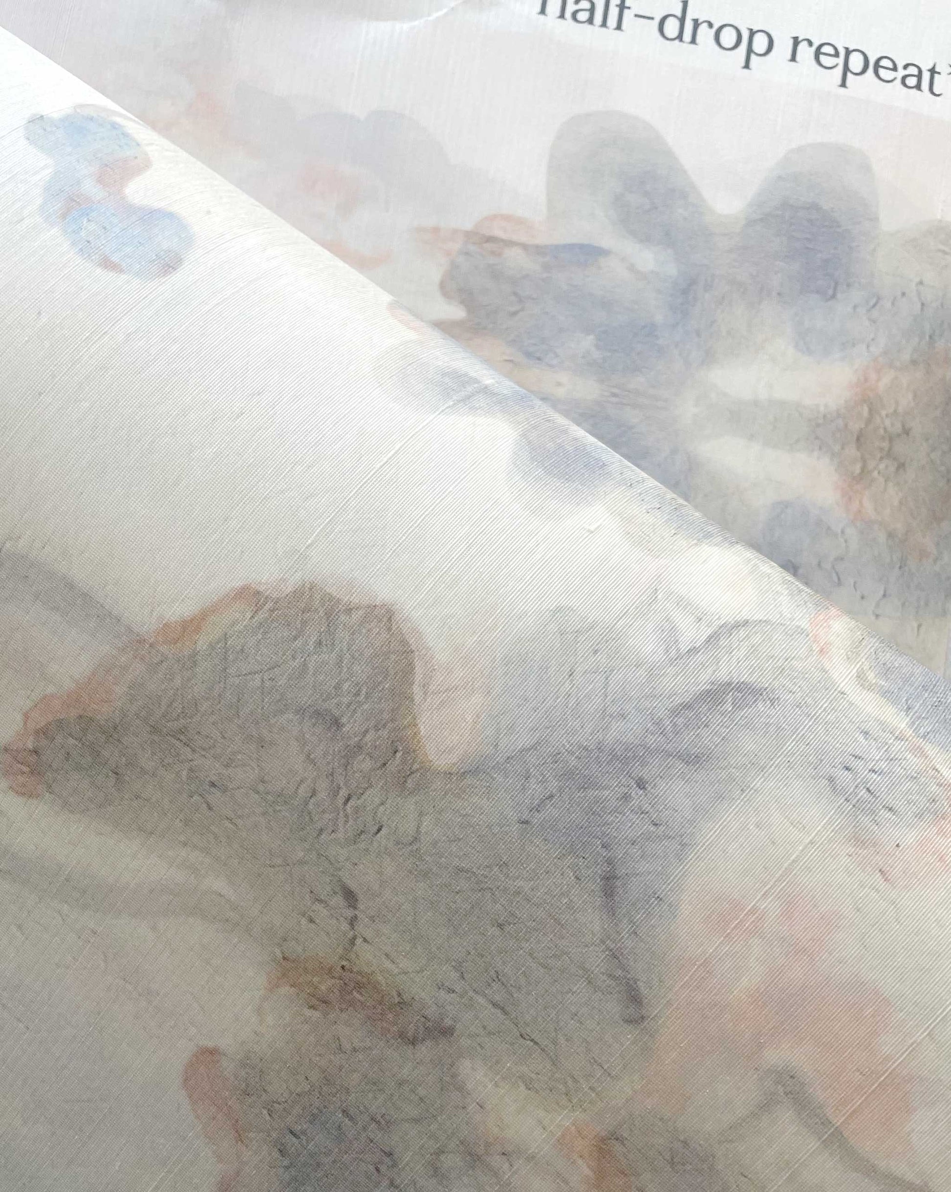 A close-up of fabric from the Clemente Silk Wallcovering||Sol, featuring a subtle, abstract watercolor pattern in muted tones reminiscent of trees, partially covering printed text that reads "half-drop repeat.
