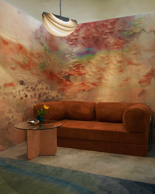 A cozy room features a rust-colored corduroy sofa, a round glass table with a flower vase, and the Empyrean Silk Mural||Sol abstract watercolor wall mural under diffused ceiling lighting, showcasing the epitome of high-end interior projects.