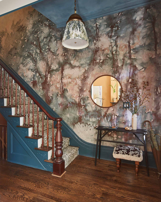 A staircase with blue trim and patterned steps is beside a wall adorned with the stunning Prospect Silk Wallcovering in Verde. A round mirror, table with decor, and a light fixture complete the space.