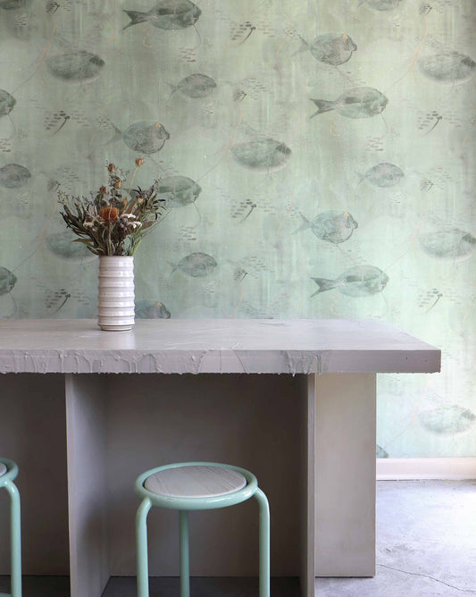 A ceramic vase with dried flowers sits on a concrete table, while a white stool with a light green frame nestles beneath it. The background features the Shoal Silk Wallcovering in Aqua, adorned with tropical fish illustrations.