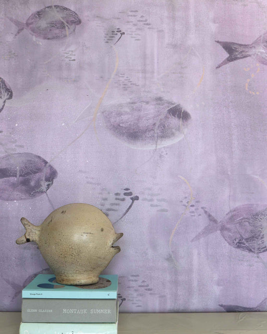 Eskayel’s Shoal wallpaper in Cay with its purple hues is printed on paper backed silk and installed in a room behind a table with a fish vase and books on top.