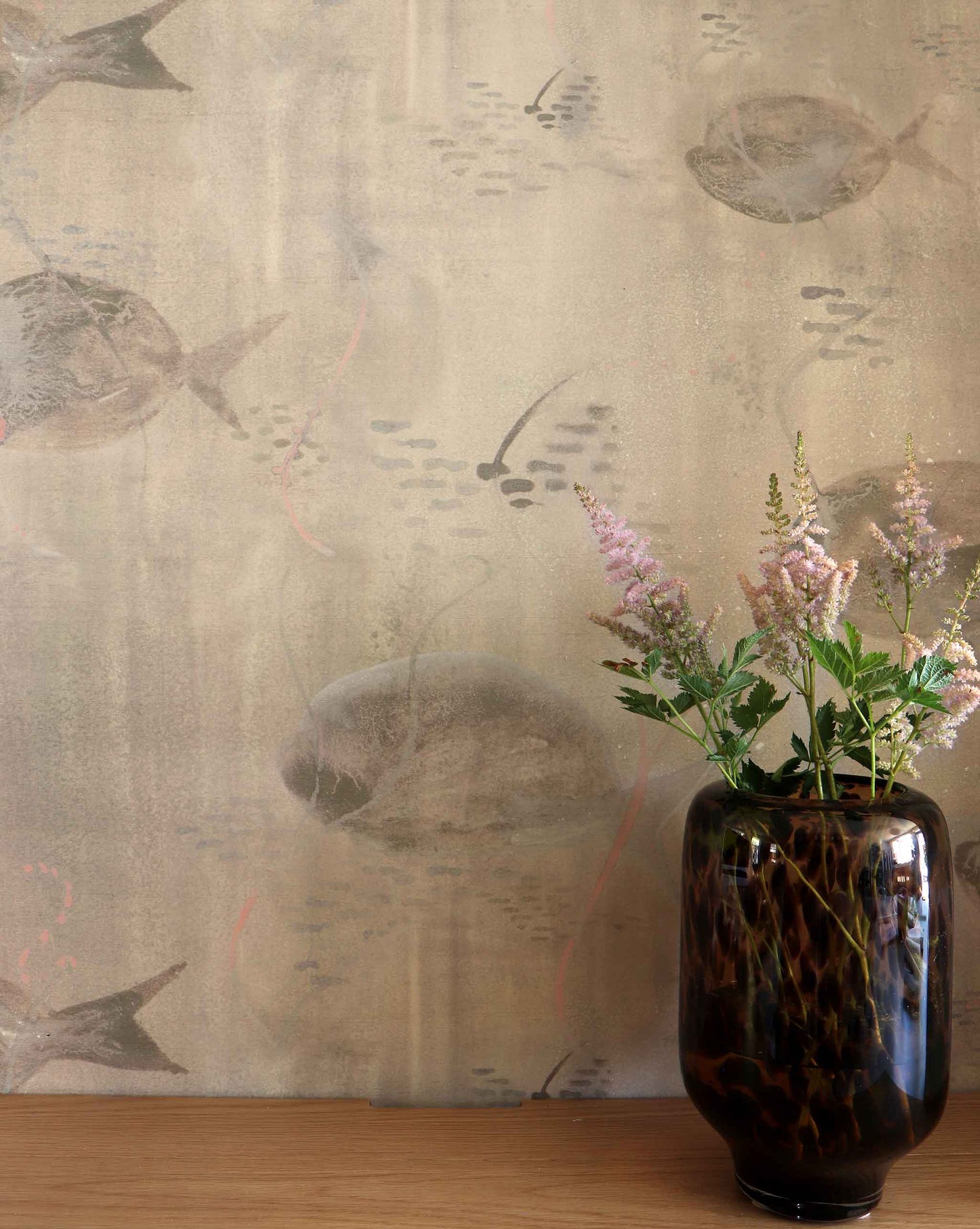 Eskayel’s Shoal wallpaper in Shell with its beige hues is printed on paper backed silk and installed in a room behind a wooden table with a vase of flowers on top.