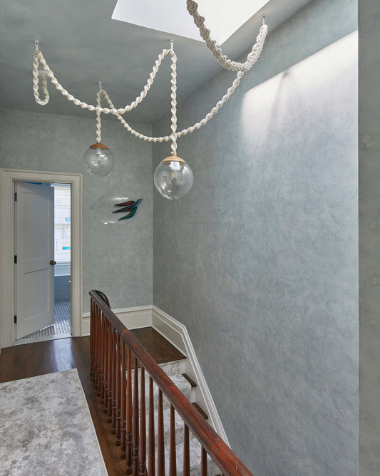 A hallway with wooden banisters, two large hanging glass light fixtures, light blue textured walls reminiscent of Stratus Silk Wallcovering in Sky, and an open doorway leading to a room with pale blue walls and a tiled floor.