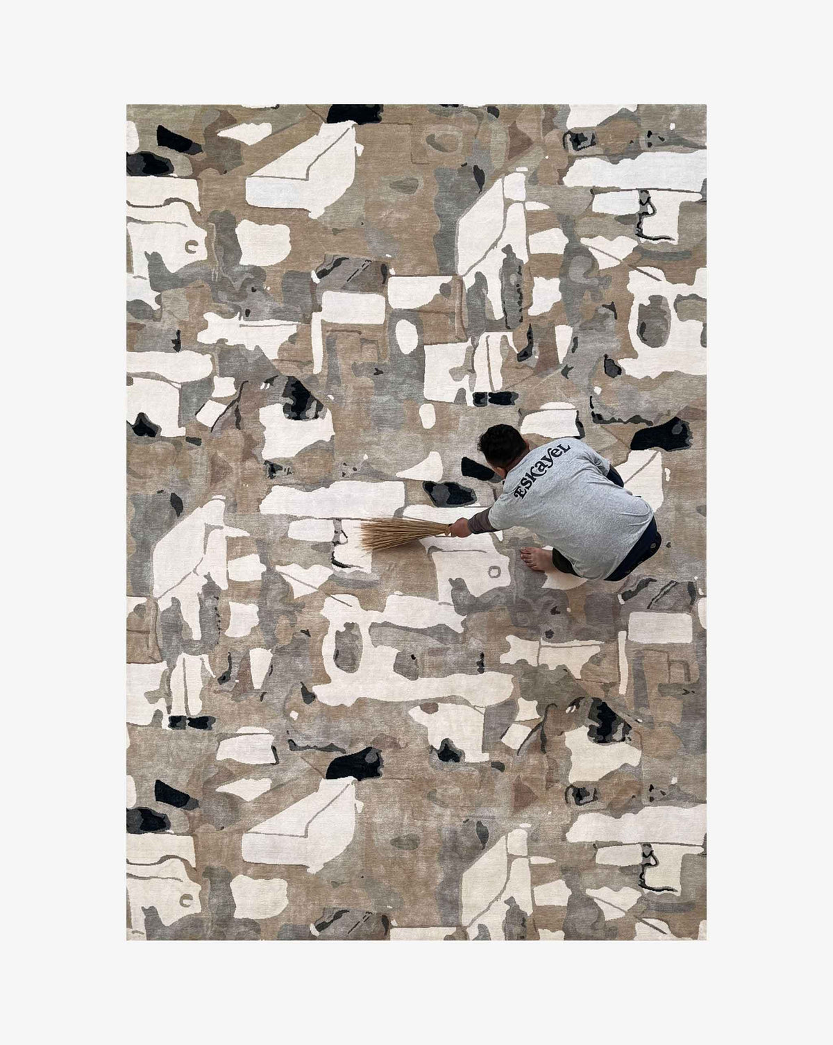 A rectangular abstract-patterned rug in beige, gray, white, and black, shown from above with a person sweeping on top of it.