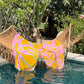 A hammock with a vibrant tropical beach towel and a colorful Plumeria Towel draped over it hangs above a swimming pool, surrounded by lush greenery.