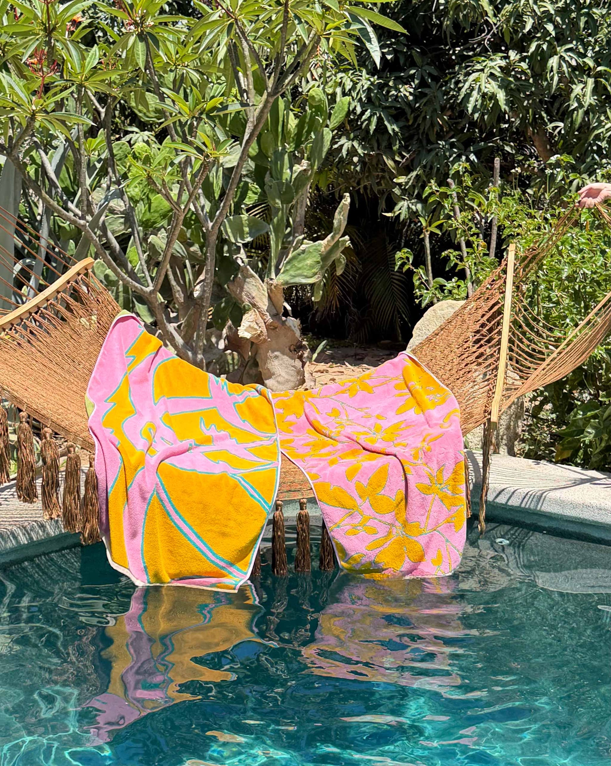 A hammock with a vibrant tropical beach towel and a colorful Plumeria Towel draped over it hangs above a swimming pool, surrounded by lush greenery.