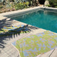 Two Plumeria Towels, including a hand-painted design, are laid out on a wooden deck beside a rectangular pool surrounded by lush plants and succulents in bright sunlight.