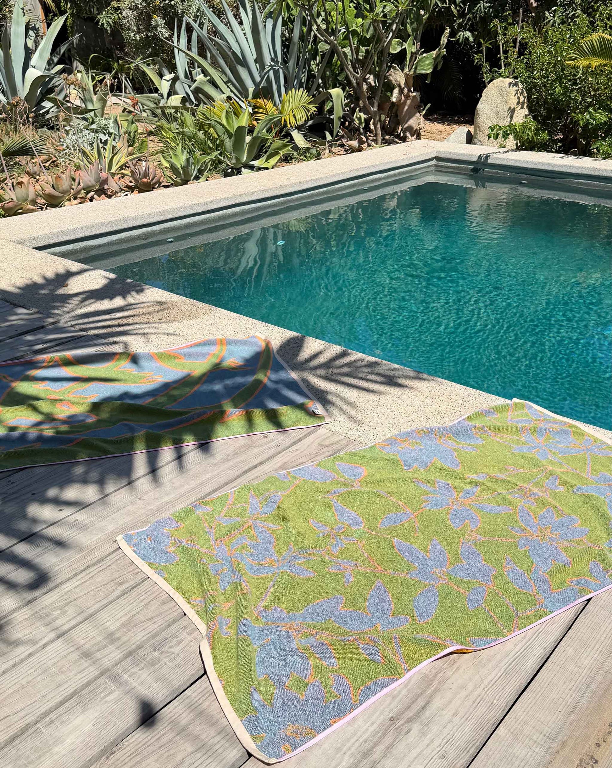 Two Plumeria Towels, including a hand-painted design, are laid out on a wooden deck beside a rectangular pool surrounded by lush plants and succulents in bright sunlight.