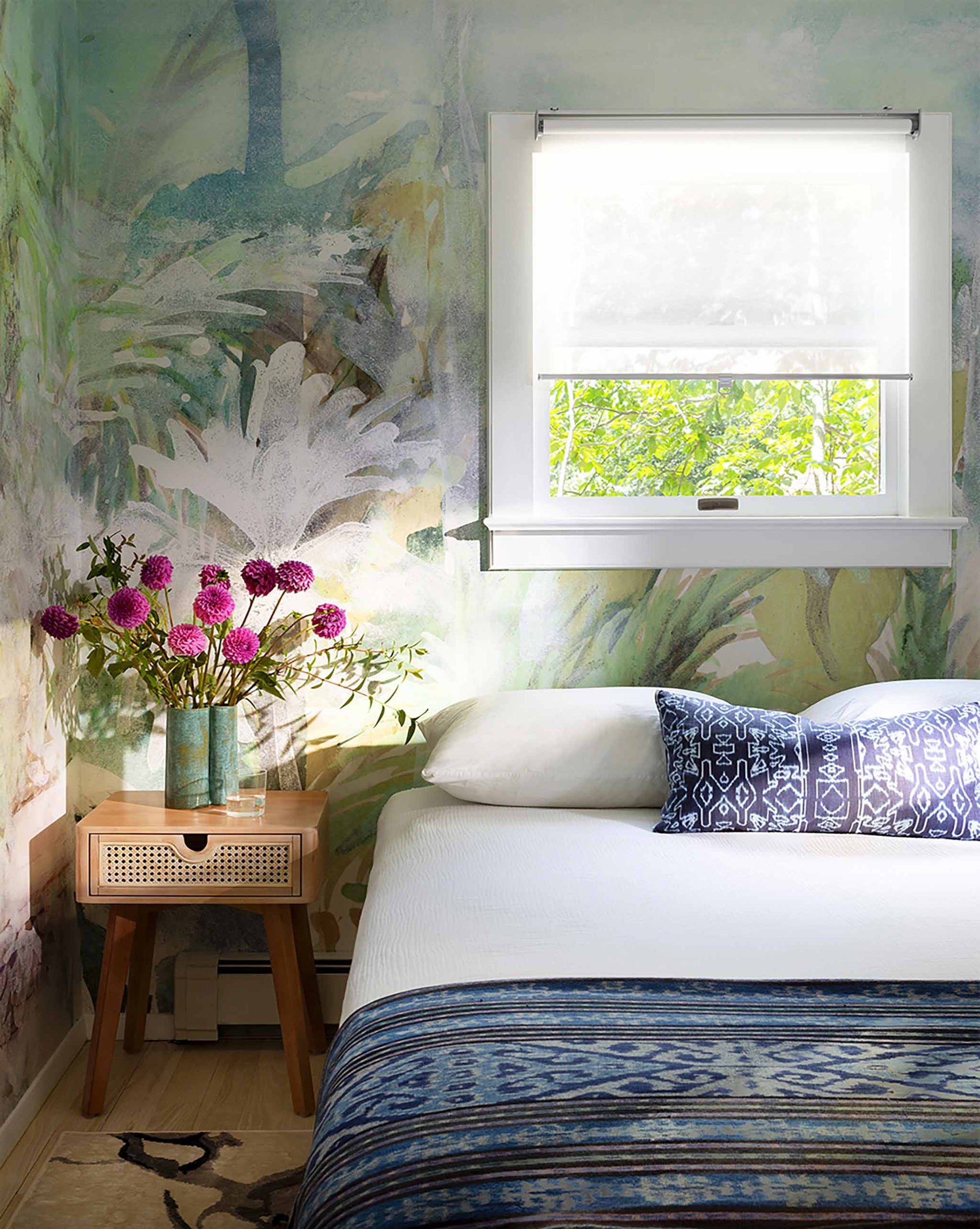 A cozy bedroom with a Regalo di Dio Wallpaper Mural||Verde, featuring a single bed with blue patterned linens, a small wooden bedside table, a vase with pink flowers, and a window showing green foliage.
