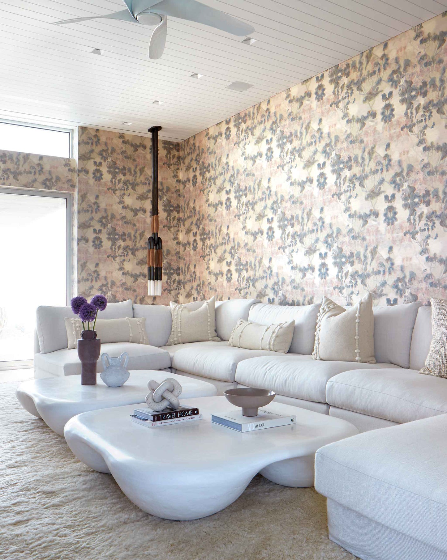 Modern living room with Clemente Wallpaper||Sol, a large white sectional sofa, unique white coffee tables, decorative books, and a hanging pendant light.