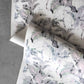 Two sheets of Cocos Wallpaper||Cay, featuring a watercolor design with pastel feathers and abstract shapes, are partially unrolled on a gray surface, evoking the elegance of luxury wallpapers.