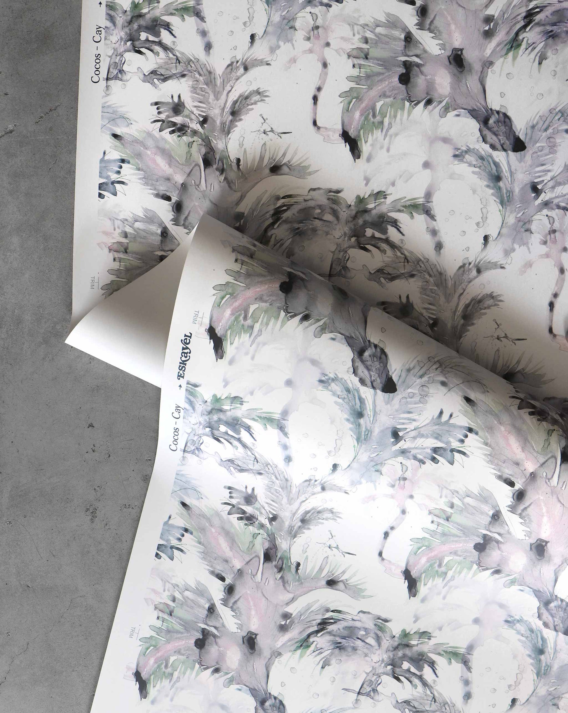 Two sheets of Cocos Wallpaper||Cay, featuring a watercolor design with pastel feathers and abstract shapes, are partially unrolled on a gray surface, evoking the elegance of luxury wallpapers.