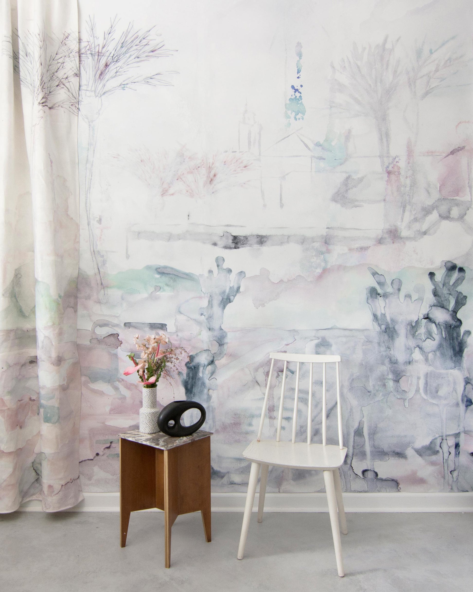 A minimalist room with a white chair, a small wooden side table with a vase of flowers, and a large mural artwork of the Palmeraie Fabric||Duomo cityscape on the wall.