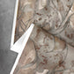A sheet of Hibiscus Lily Wallpaper||Garnet, showcasing abstract earthy watercolor patterns, is partially peeled back on a gray concrete surface.