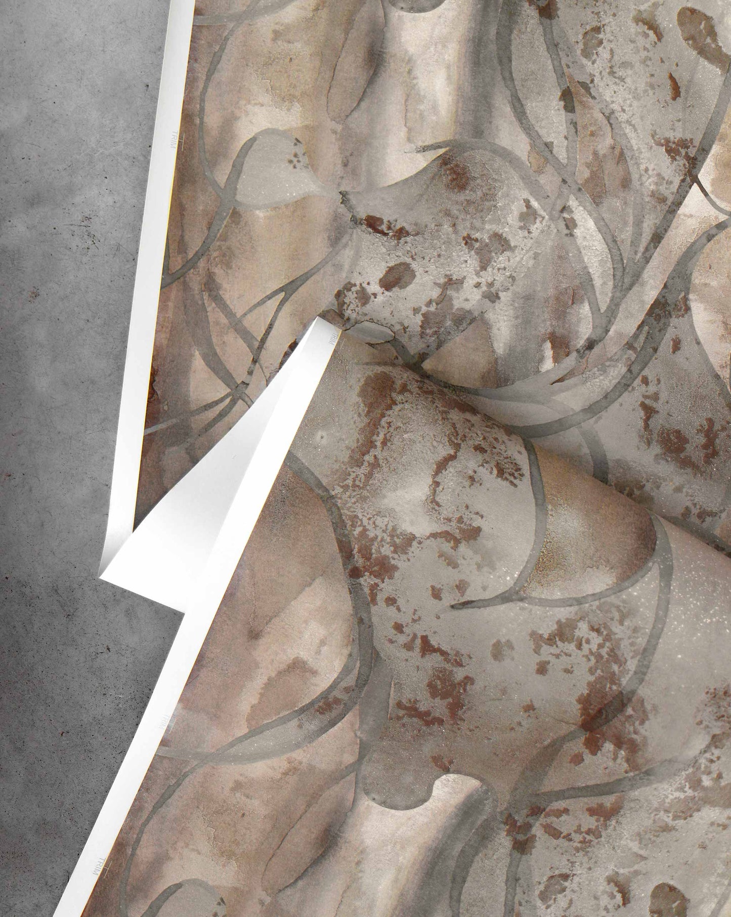 A sheet of Hibiscus Lily Wallpaper||Garnet, showcasing abstract earthy watercolor patterns, is partially peeled back on a gray concrete surface.
