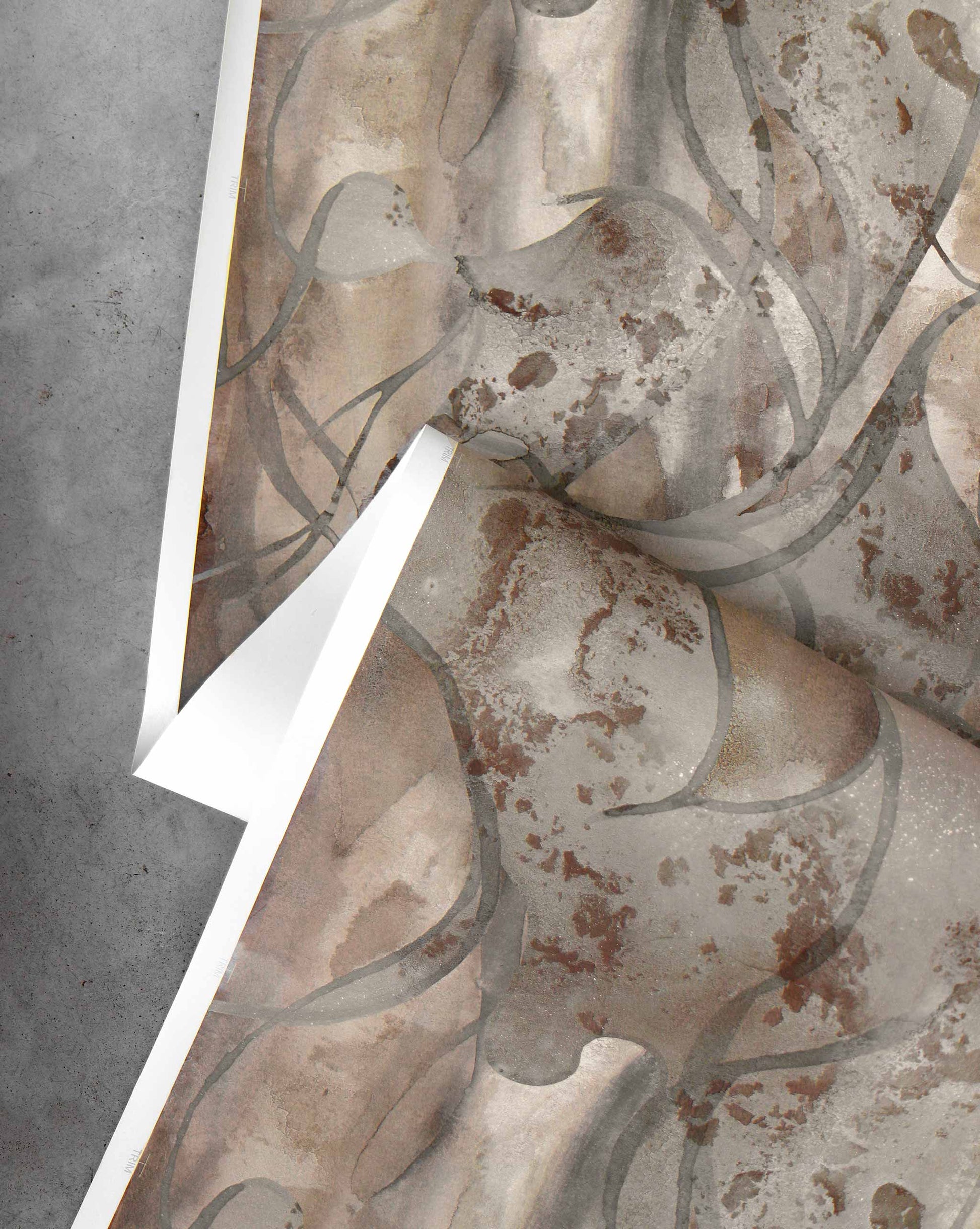 A sheet of Hibiscus Lily Wallpaper||Garnet, showcasing abstract earthy watercolor patterns, is partially peeled back on a gray concrete surface.