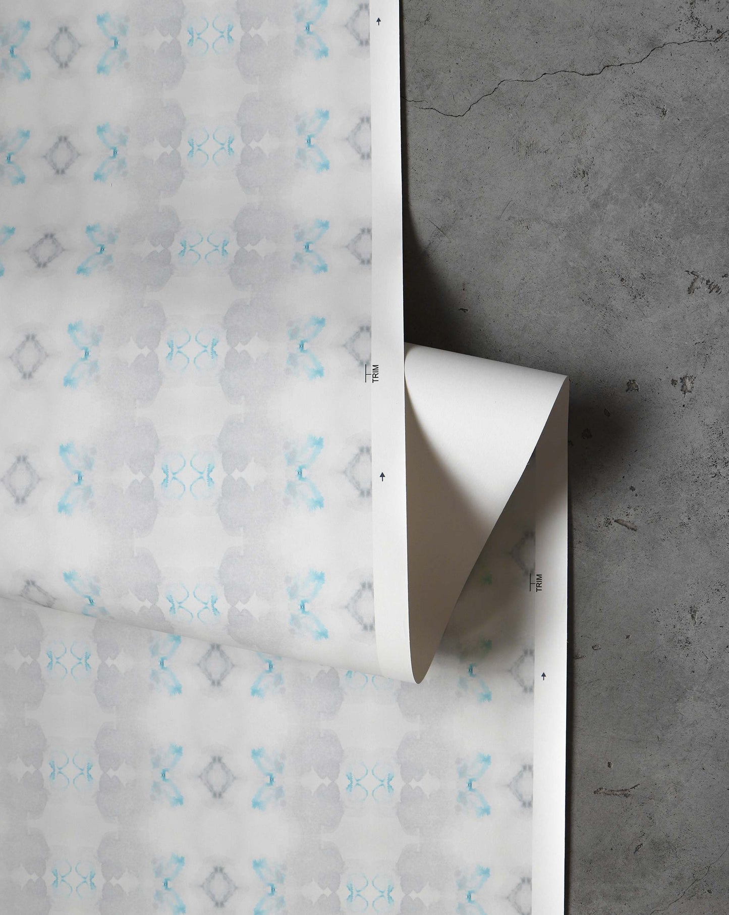 An unrolled roll of Icelandic Mist Wallpaper in Sea Green lies on a concrete floor, featuring a subtle geometric pattern with blue and gray accents.