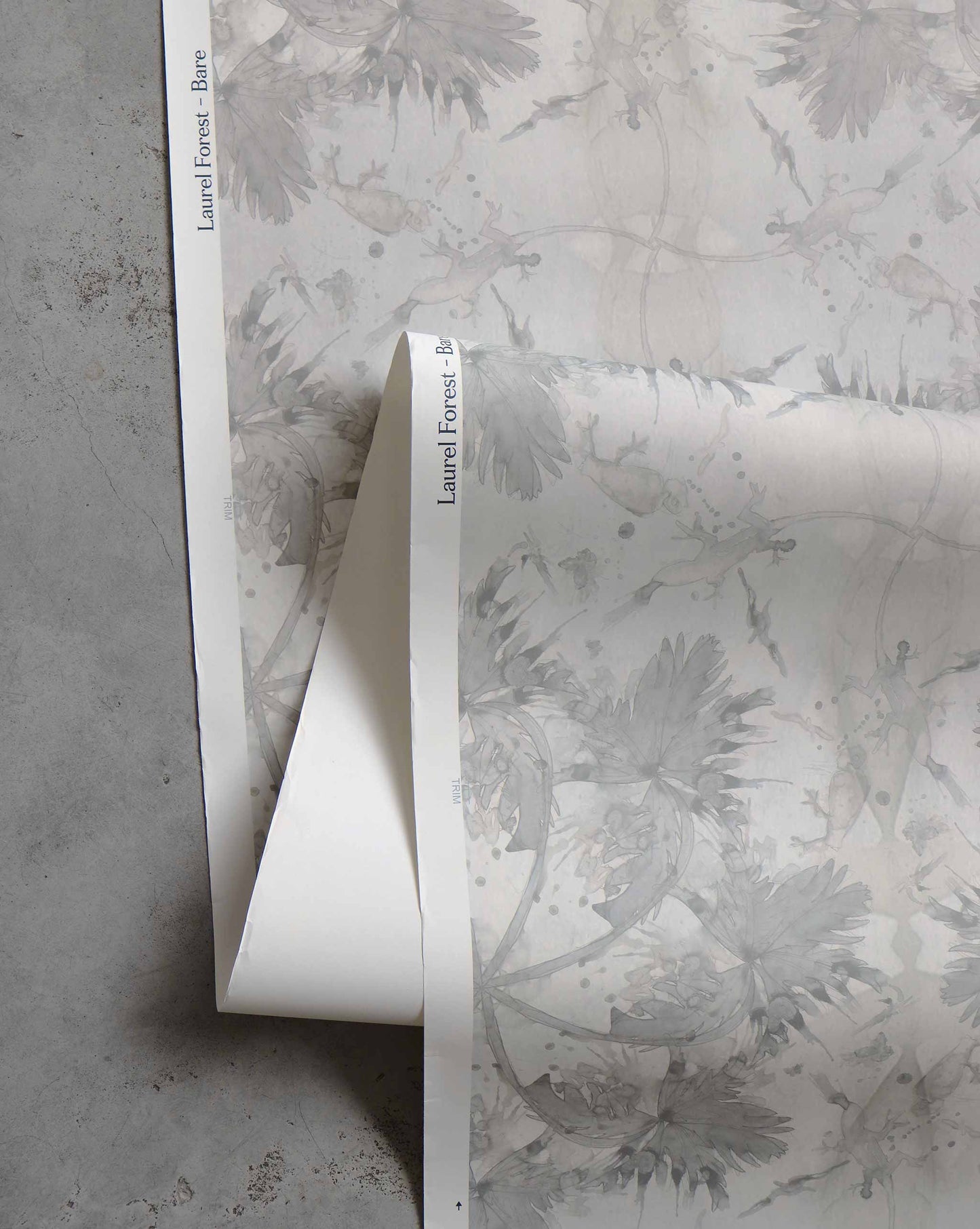 A close-up view of the high-end Laurel Forest Wallpaper in the "Bare" design, showcasing a subtle floral pattern in light grey tones on a light background, laid out on a grey surface. The luxurious fabric features a printed edge label "Laurel Forest, Bone.
