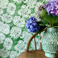Margherita wallpaper with a floral lace pattern on a green background, featuring a vase with purple flowers.