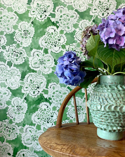 Margherita wallpaper with a floral lace pattern on a green background, featuring a vase with purple flowers.