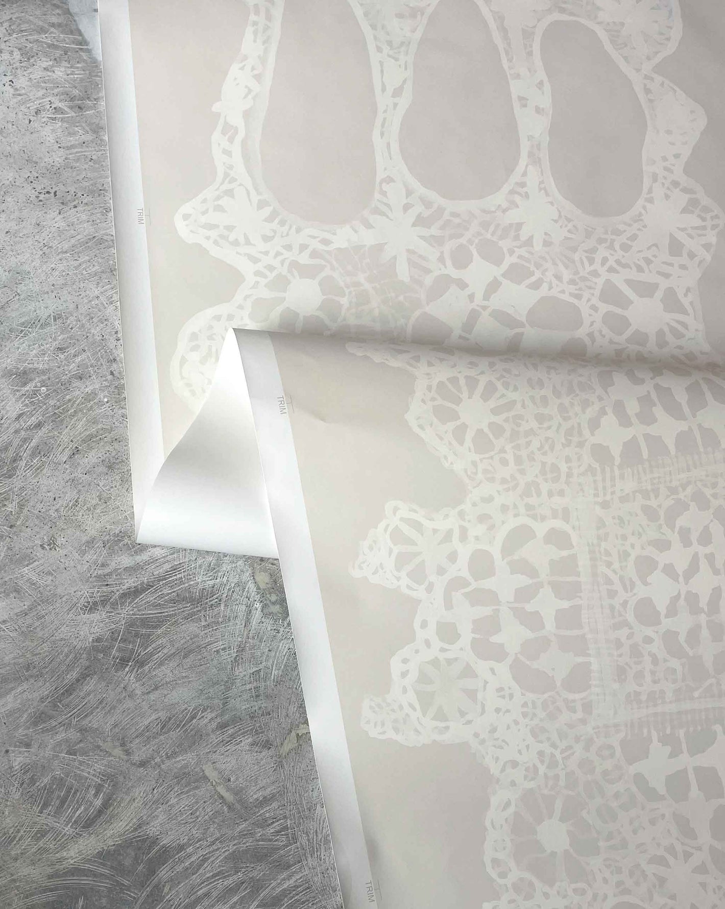 Close-up of a neutral lace wallpaper in Eskayel's Nonna's Lace Ricotta pattern on a textured gray surface.