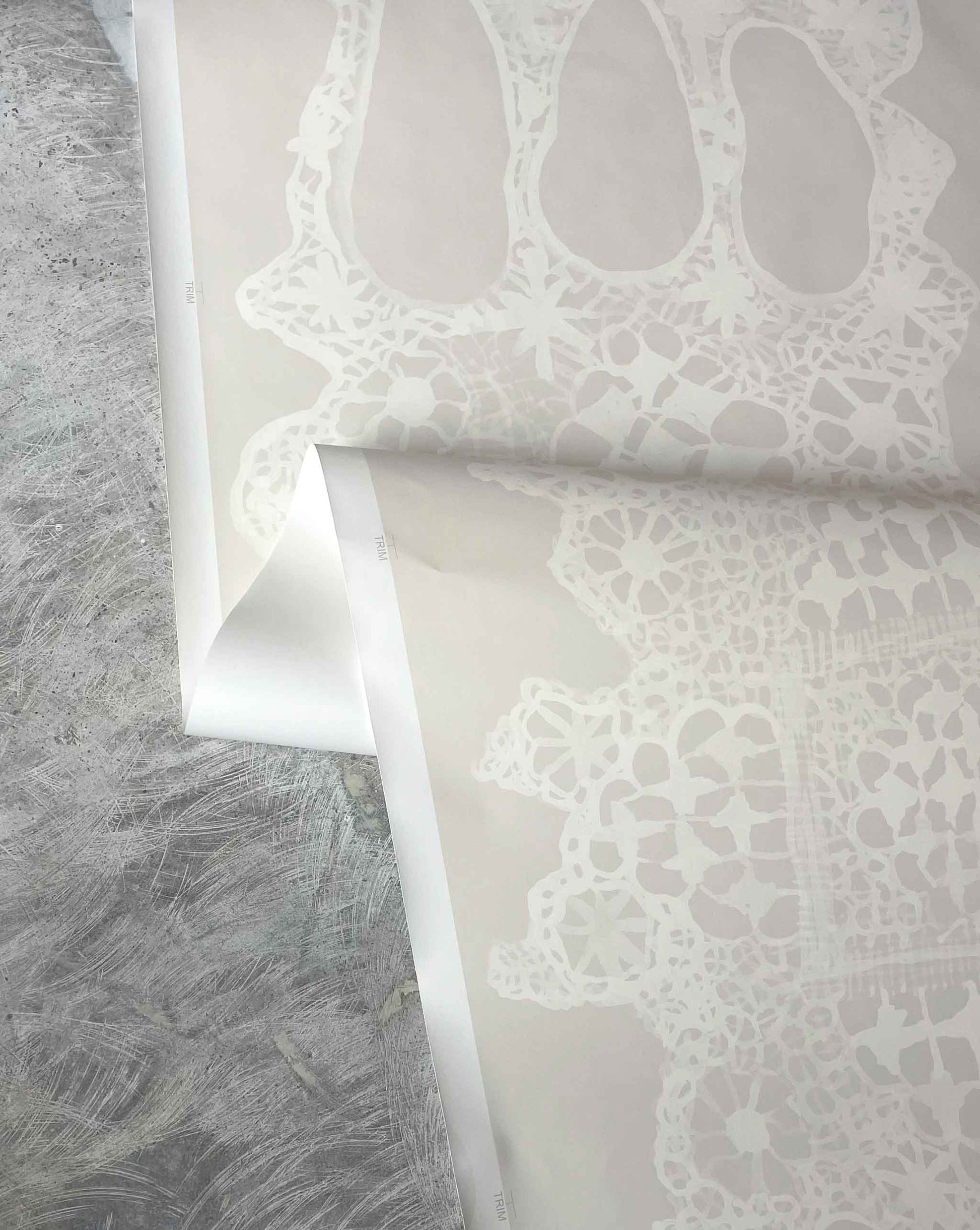 Close-up of a neutral lace wallpaper in Eskayel's Nonna's Lace Ricotta pattern on a textured gray surface.