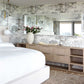 A modern bedroom in Presidio features a large bed with white linens, a dresser with a large mirror above, a vase of pink flowers, and neutral-toned decor. The room is enhanced by high-end Sol wallpaper from Presidio and luxury fabric accents, including a textured rug to complete the look.