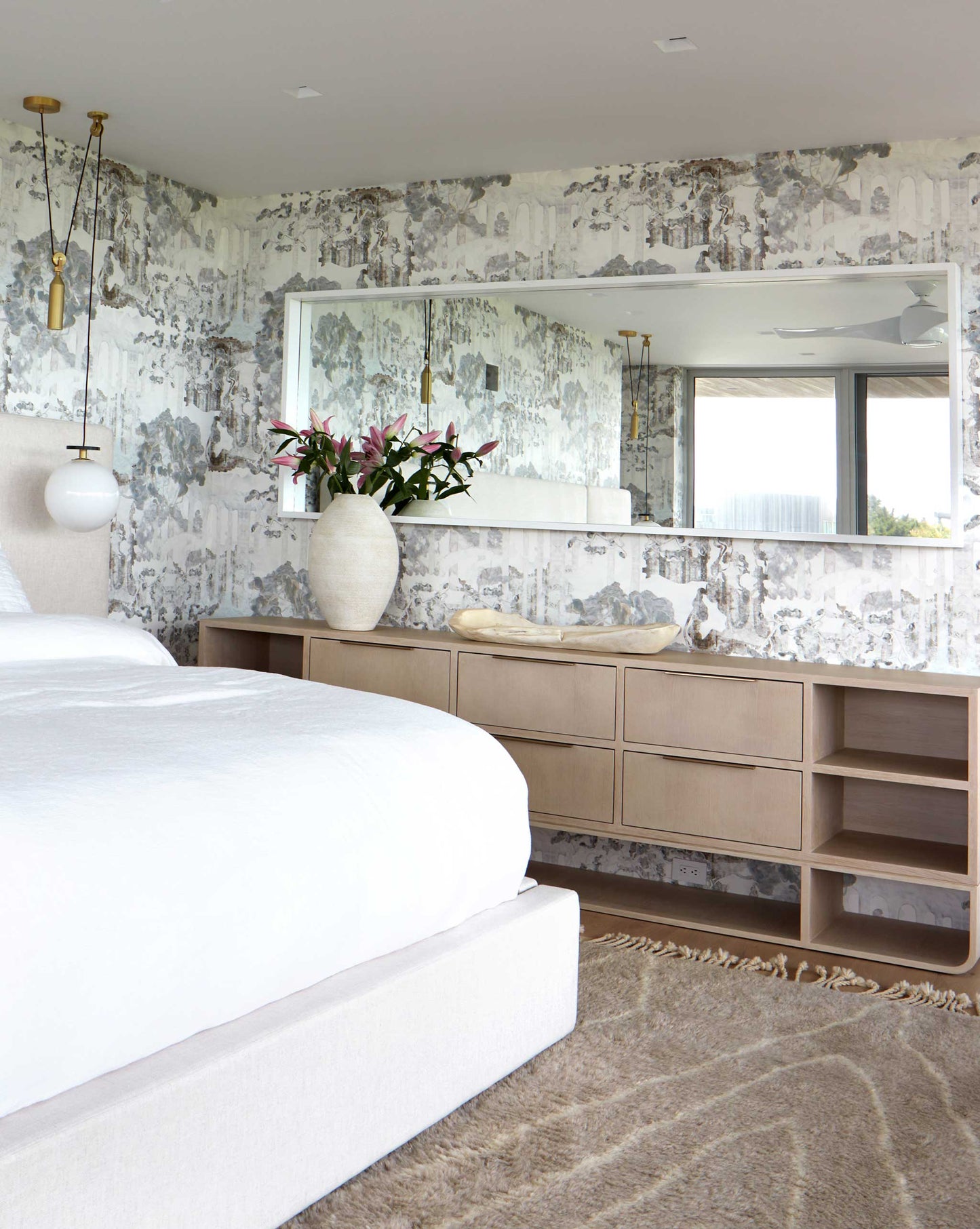 A modern bedroom in Presidio features a large bed with white linens, a dresser with a large mirror above, a vase of pink flowers, and neutral-toned decor. The room is enhanced by high-end Sol wallpaper from Presidio and luxury fabric accents, including a textured rug to complete the look.