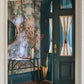 Entryway with a teal door, sheer curtain panels, and Prospect Wallpaper Mural in Verde featuring a round mirror. A table holds a vase with branches and a tray, while an umbrella stands nearby.