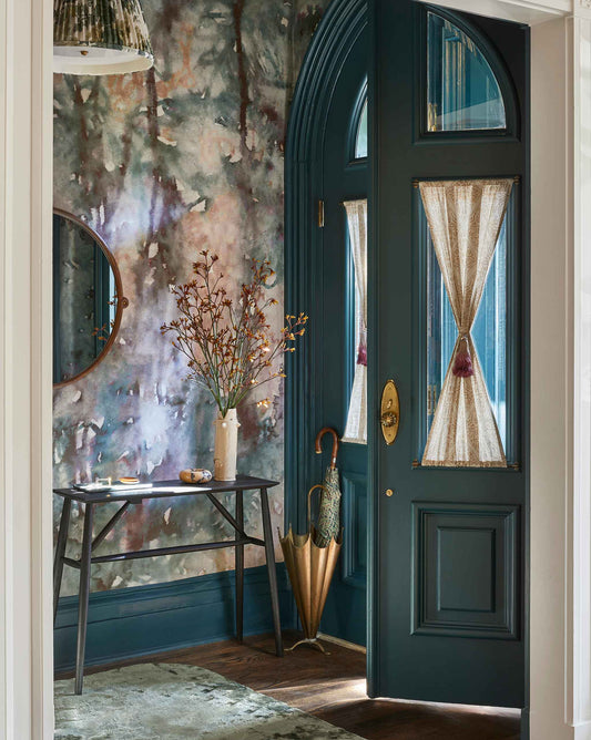 Entryway with a teal door, sheer curtain panels, and Prospect Wallpaper Mural in Verde featuring a round mirror. A table holds a vase with branches and a tray, while an umbrella stands nearby.