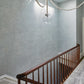 A wooden staircase with a railing descends alongside a textured light blue wall adorned with Stratus Wallpaper in the Sky pattern. Two hanging glass globe lights are visible above.