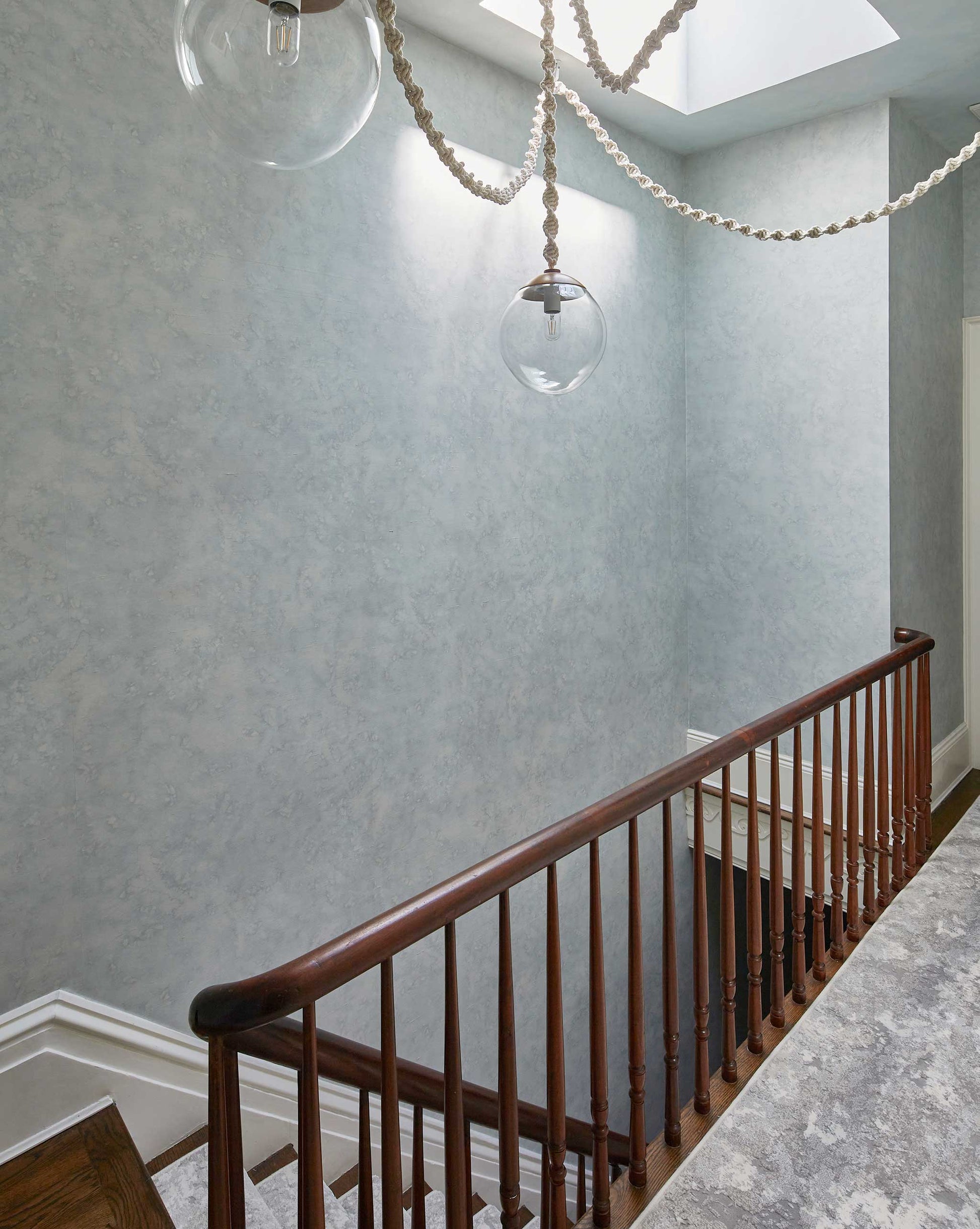 A wooden staircase with a railing descends alongside a textured light blue wall adorned with Stratus Wallpaper in the Sky pattern. Two hanging glass globe lights are visible above.