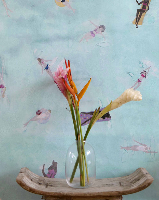 Eskayel's Swim wallpaper in the colorway Sea in light blue with waterborne female figures installed as an accent wall with a vase of flowers and a wooden stool.