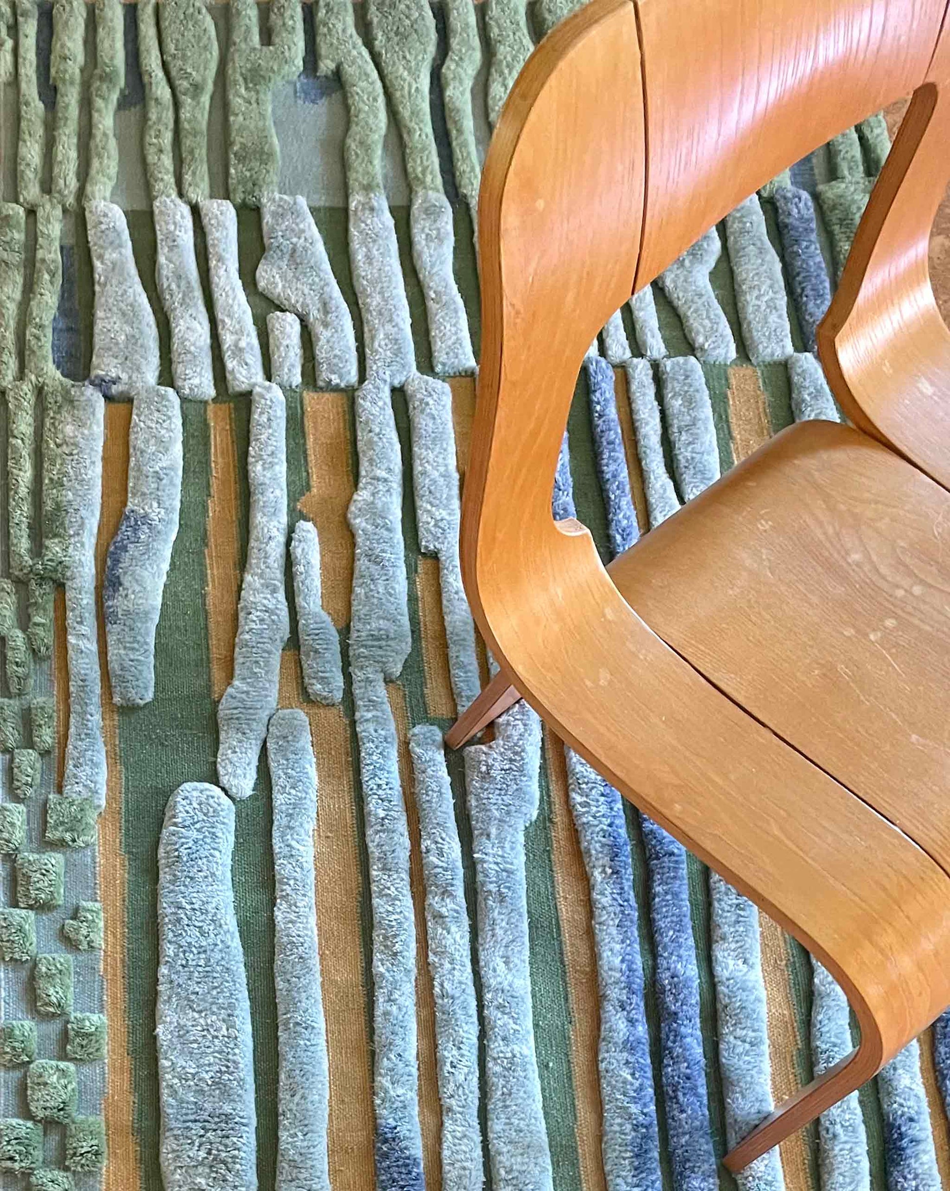 A curved wooden chair sits on our studio-designed Pyrus Stripe Hand Knotted Rug in Low Tide, which features textured, raised vertical blue and green stripes | Eskayel