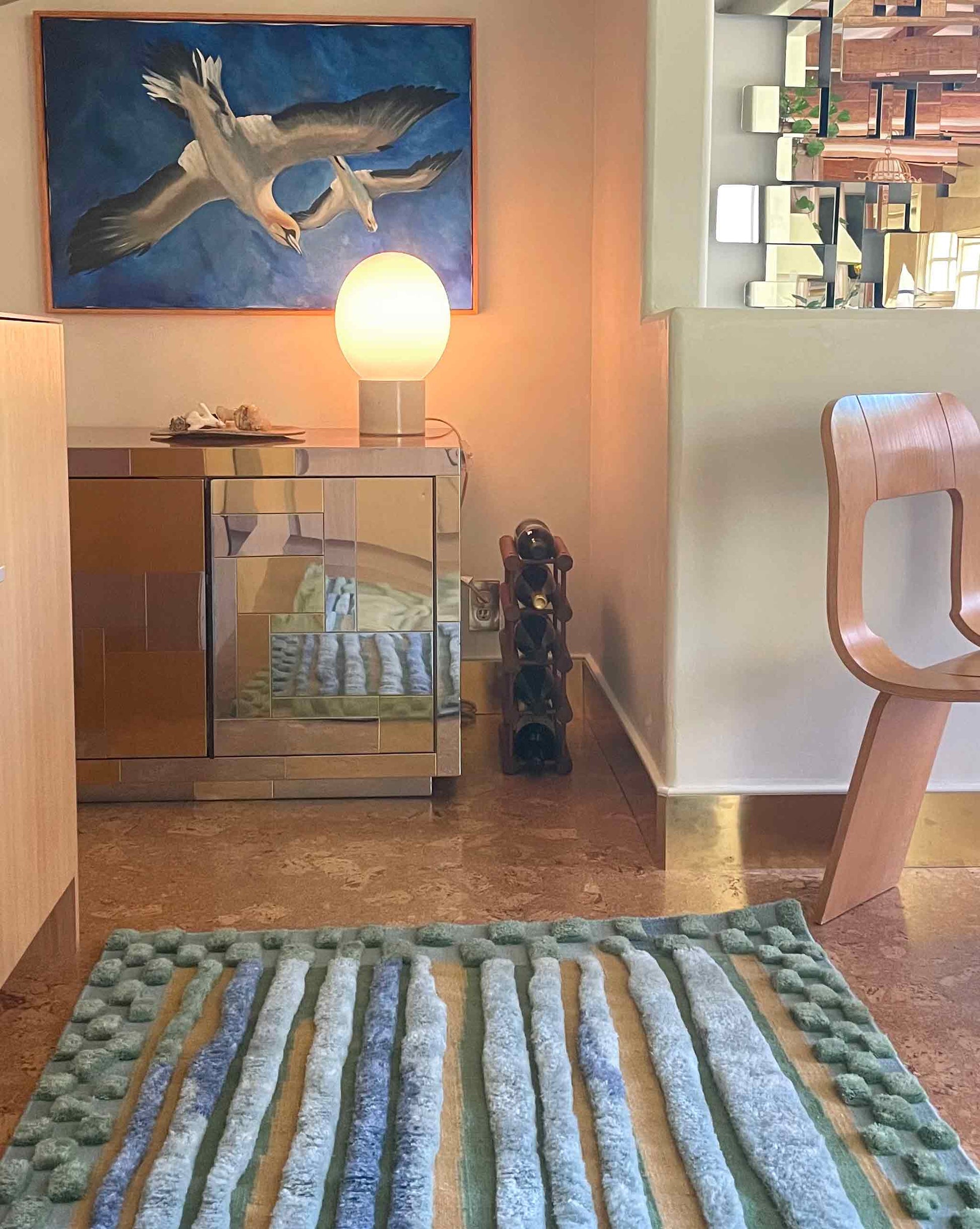 Pyrus Stripe Rug in Low Tide – hand-knotted studio-designed rug in green and blue installed in a kitchen with a mirror-front cabinet, a spherical lamp, a wine rack, a modern chair, and a painting of a flying bird on the wall | Eskayel  