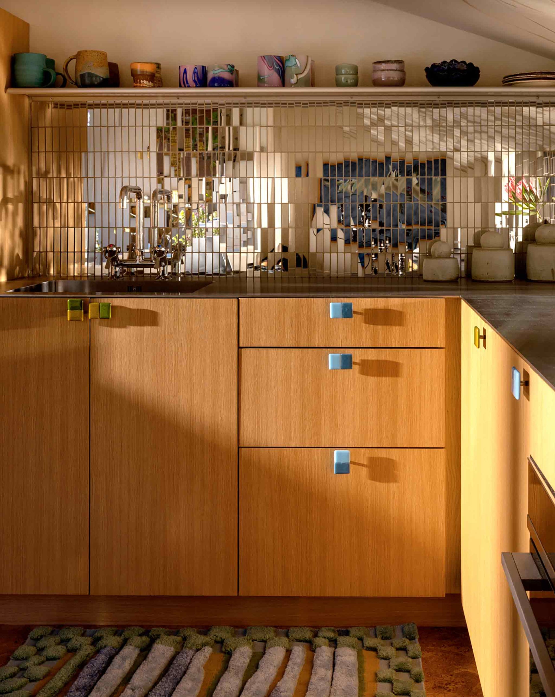 Modern kitchen with light wood cabinets, colorful knobs, a metal backsplash, open shelves of ceramics, and our studio-designed Pyrus Stripe Hand Knotted Rug in the colorway  Low Tide on the floor. Natural light casts soft shadows across the surfaces | Eskayel