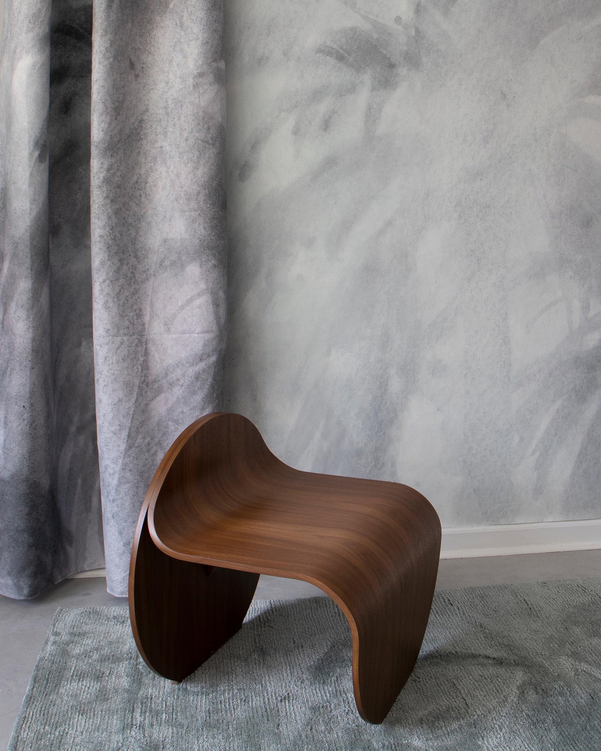 A wooden stool is sitting on a Reflettere Fabric Aqua rug in front of a mural wallpaper